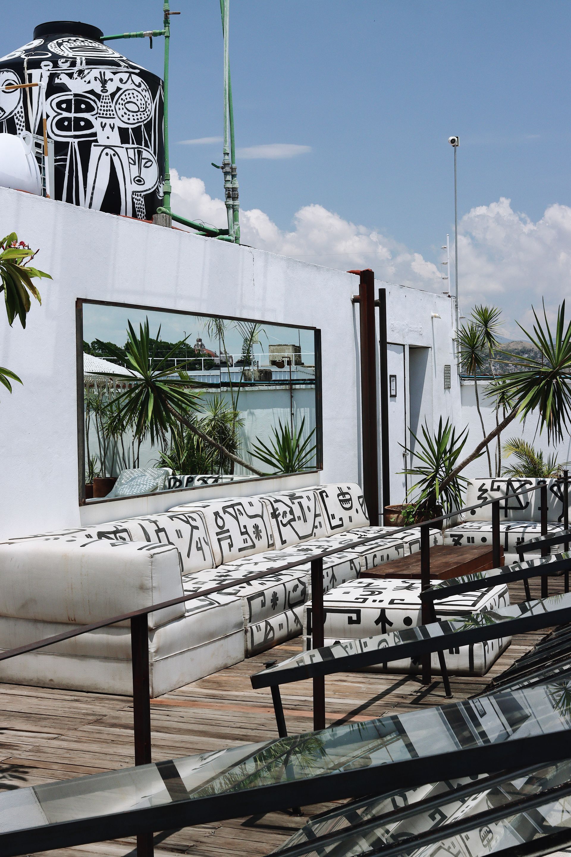 Outdoor rooftop lounge with white couches, glass tables, and palm plants under a blue sky