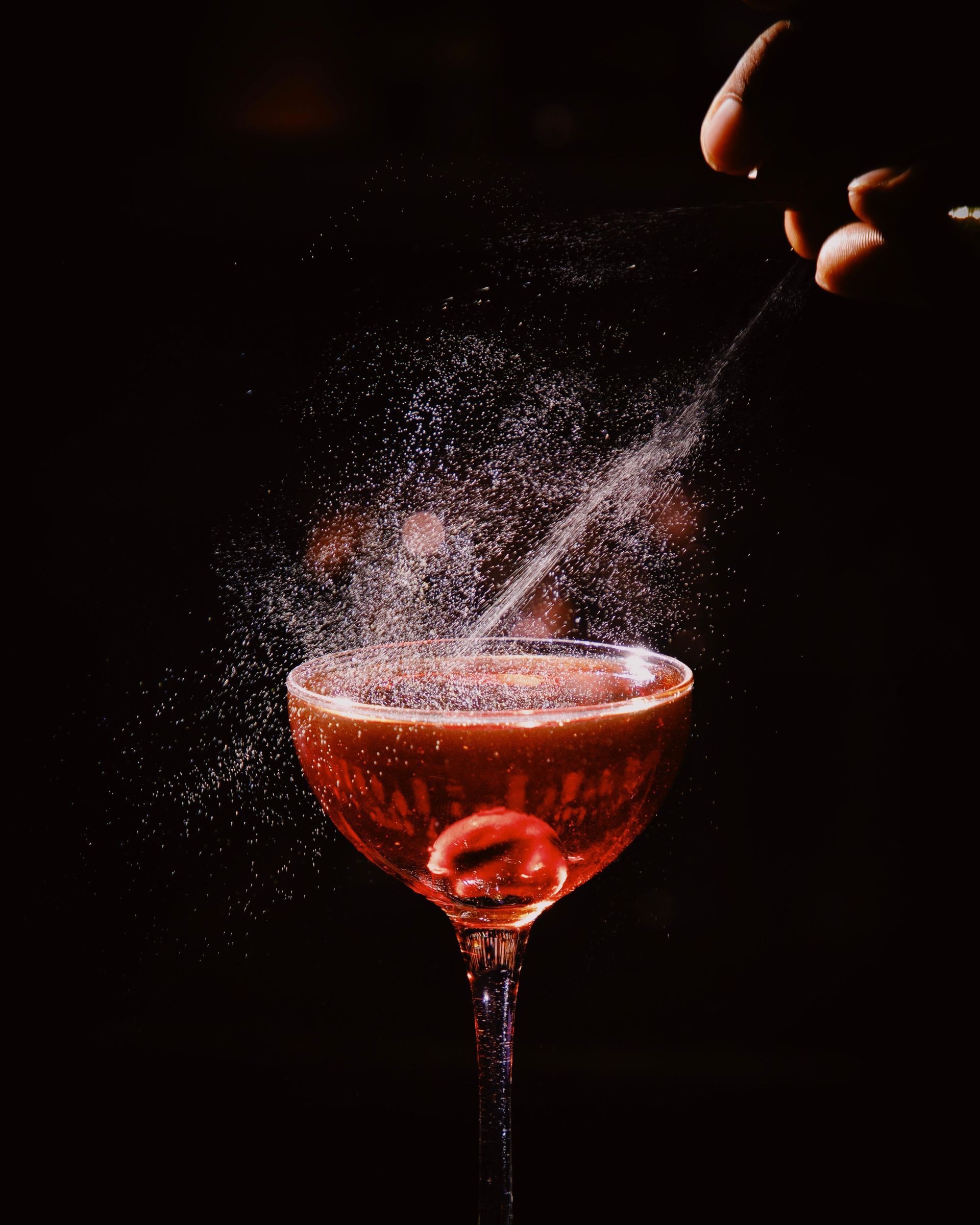 Red cocktail in a coupe glass being sprayed with a fine mist against a dark background.