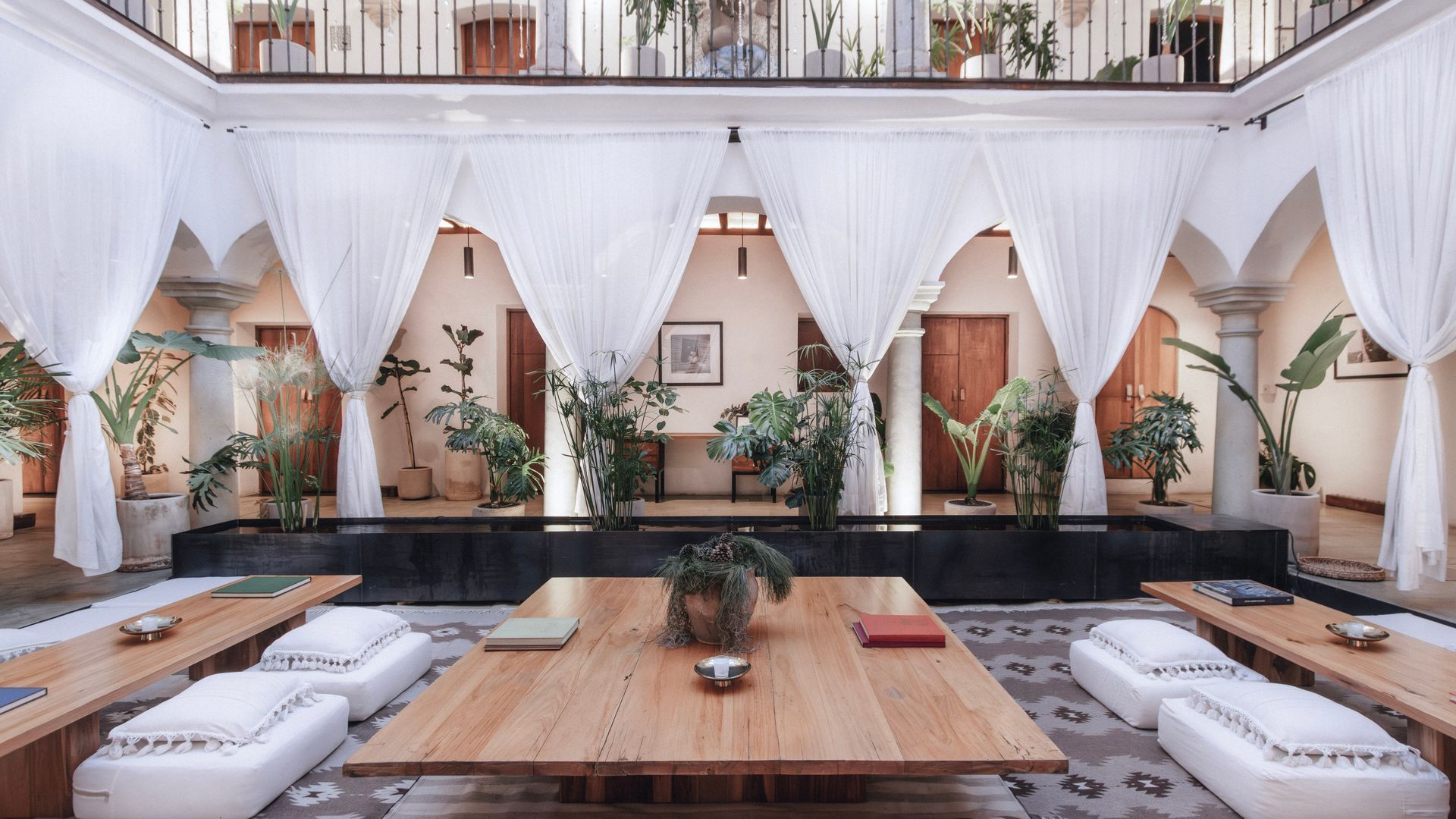 Bright, airy lounge with white drapes, wooden tables, low cushions, and potted plants around a central courtyard.