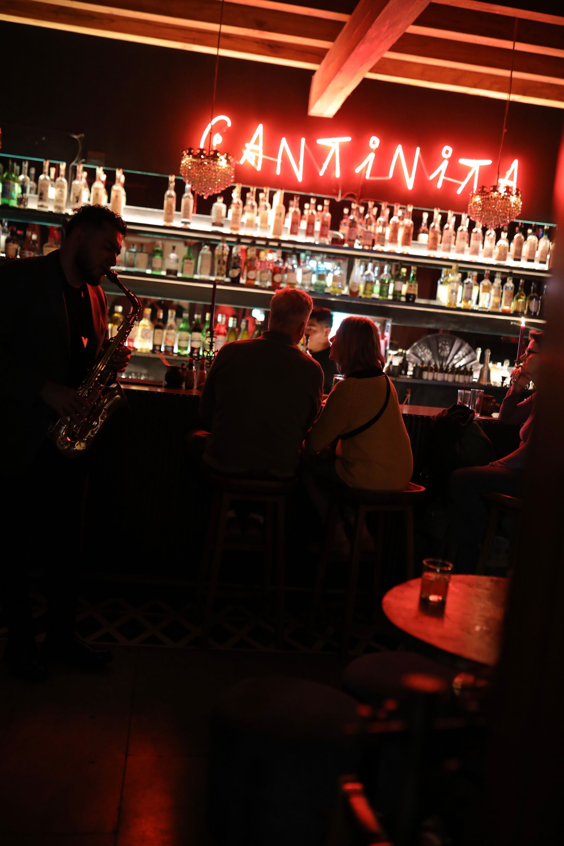 Dimly lit bar interior with a red neon “CANTINITA” sign, shelves of bottles, and patrons seated at the counter