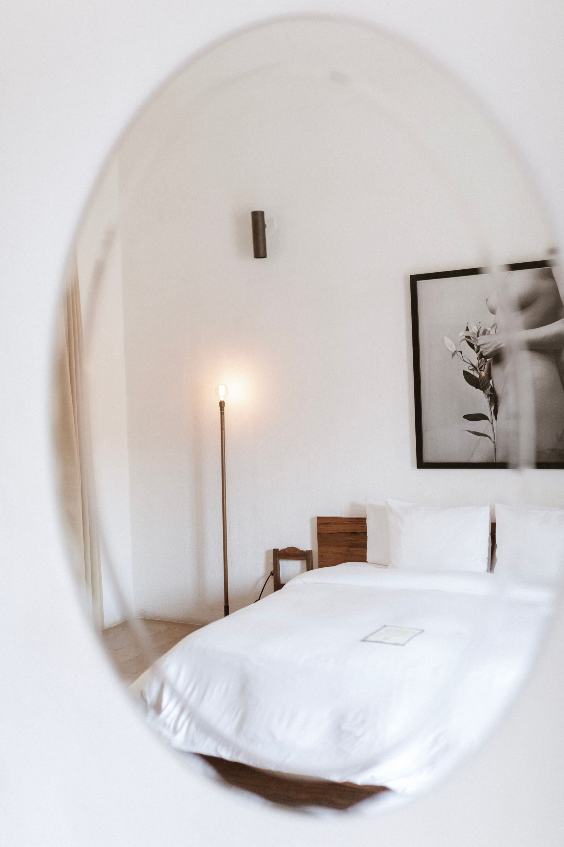 Minimal bedroom with white bedding, tall lamp, wooden chair, and wall art in an oval mirror.