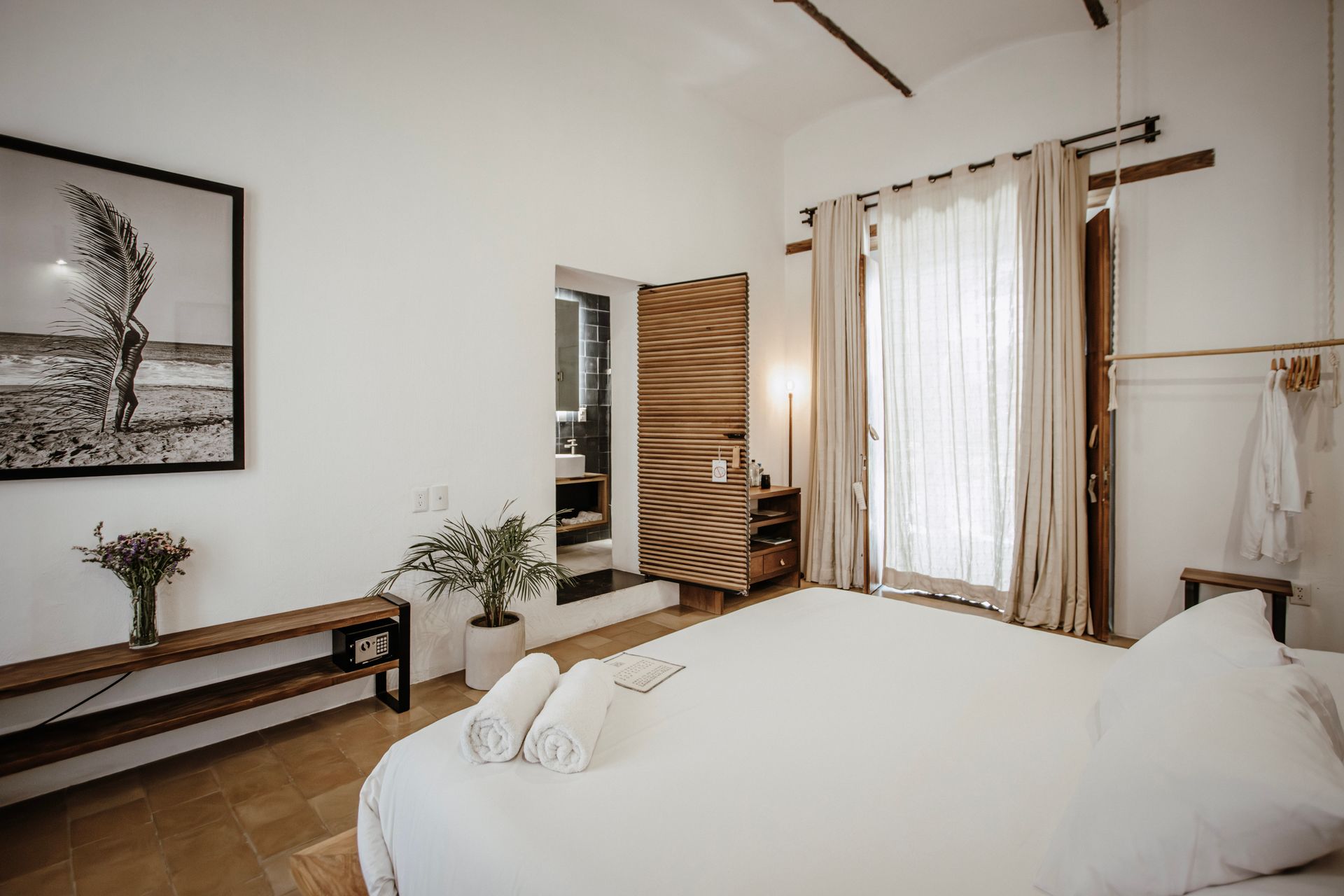 Bright bedroom with white bedding, wooden furniture, wall art, and sheer curtains by a window.