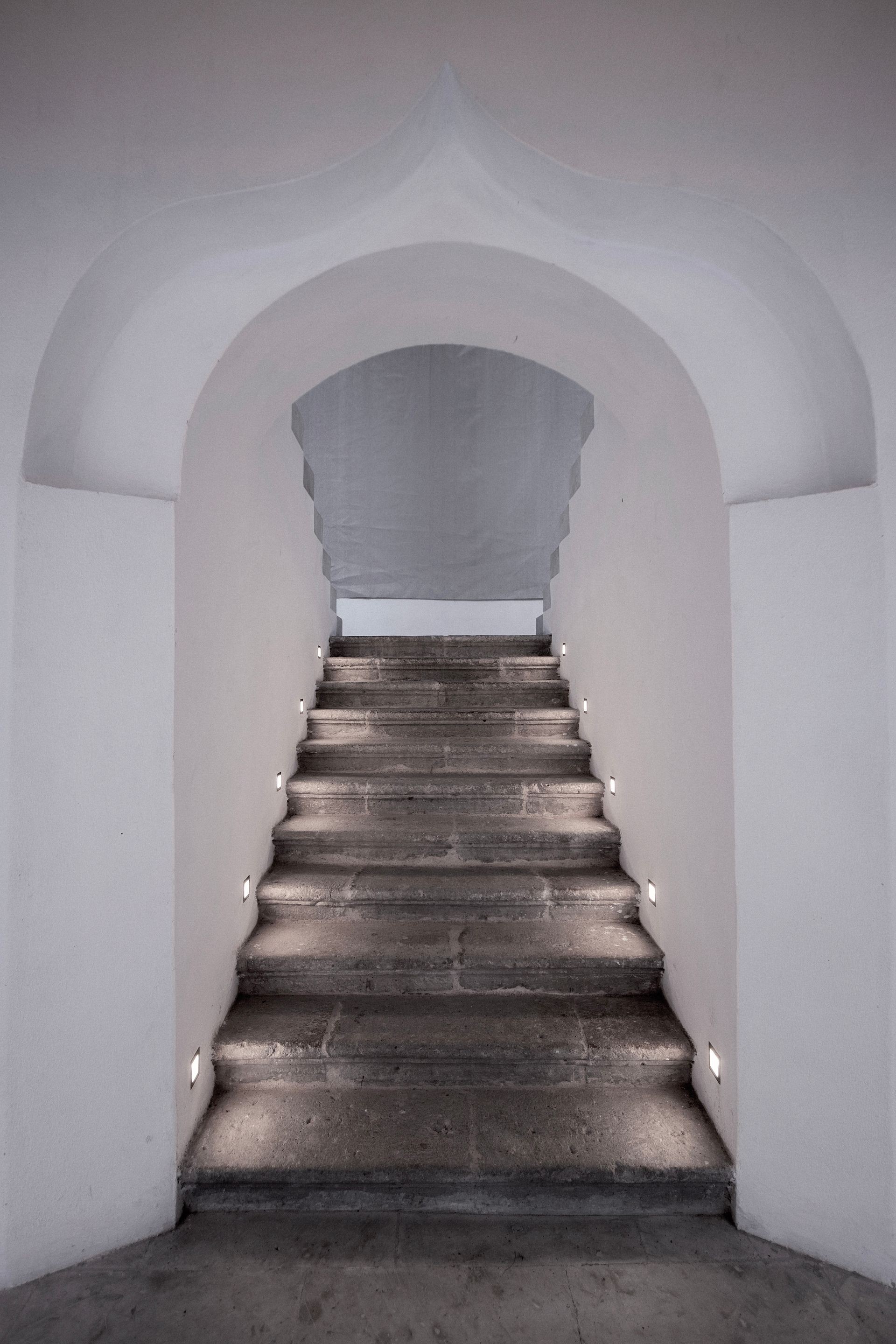 Narrow stone stairway under a white archway leading upward in a dim interior corridor