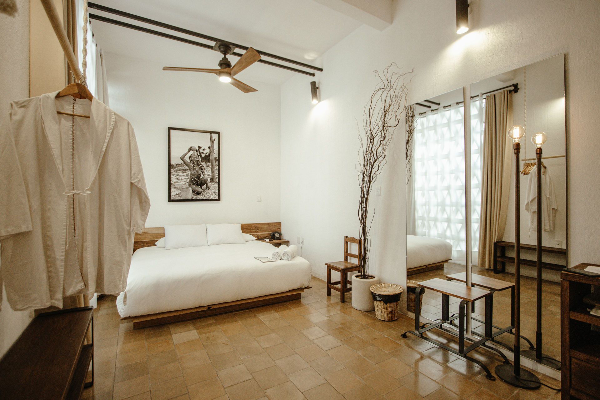 Minimalist beige bedroom with a low bed, hanging shirts, and a window beside a small desk.