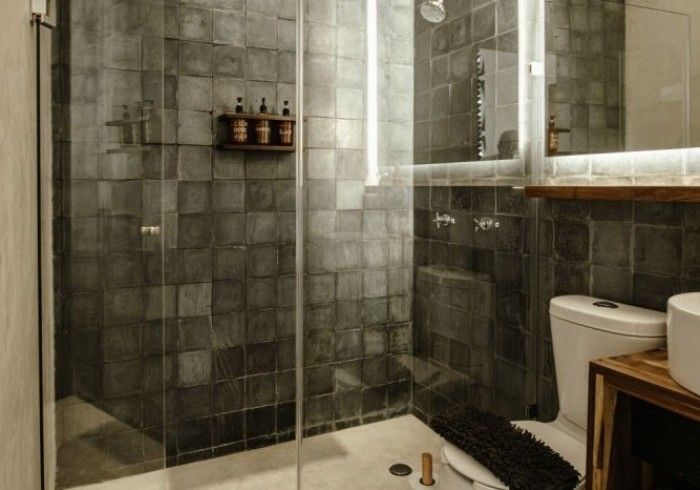 Small tiled bathroom with glass shower, toilet, sink, and wooden shelf.