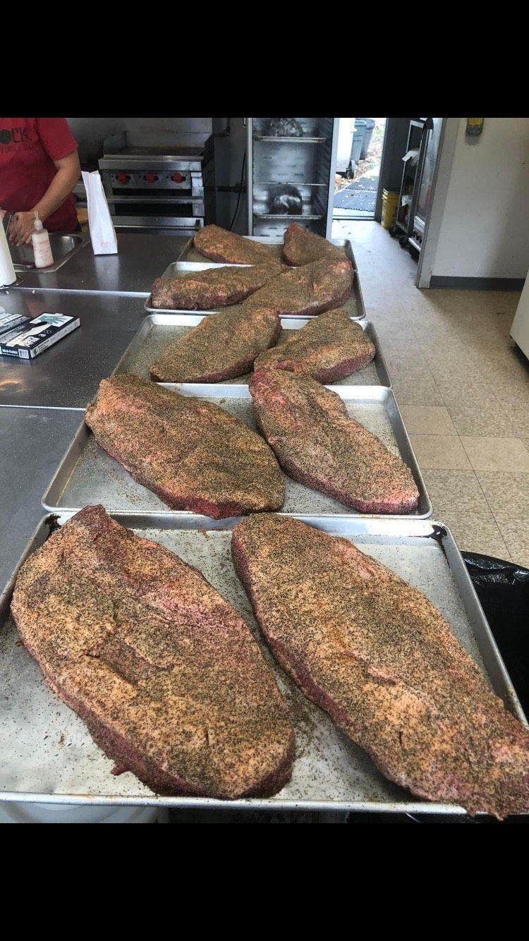 Seasoned beef briskets arranged in rows on metal baking sheets in a commercial kitchen.