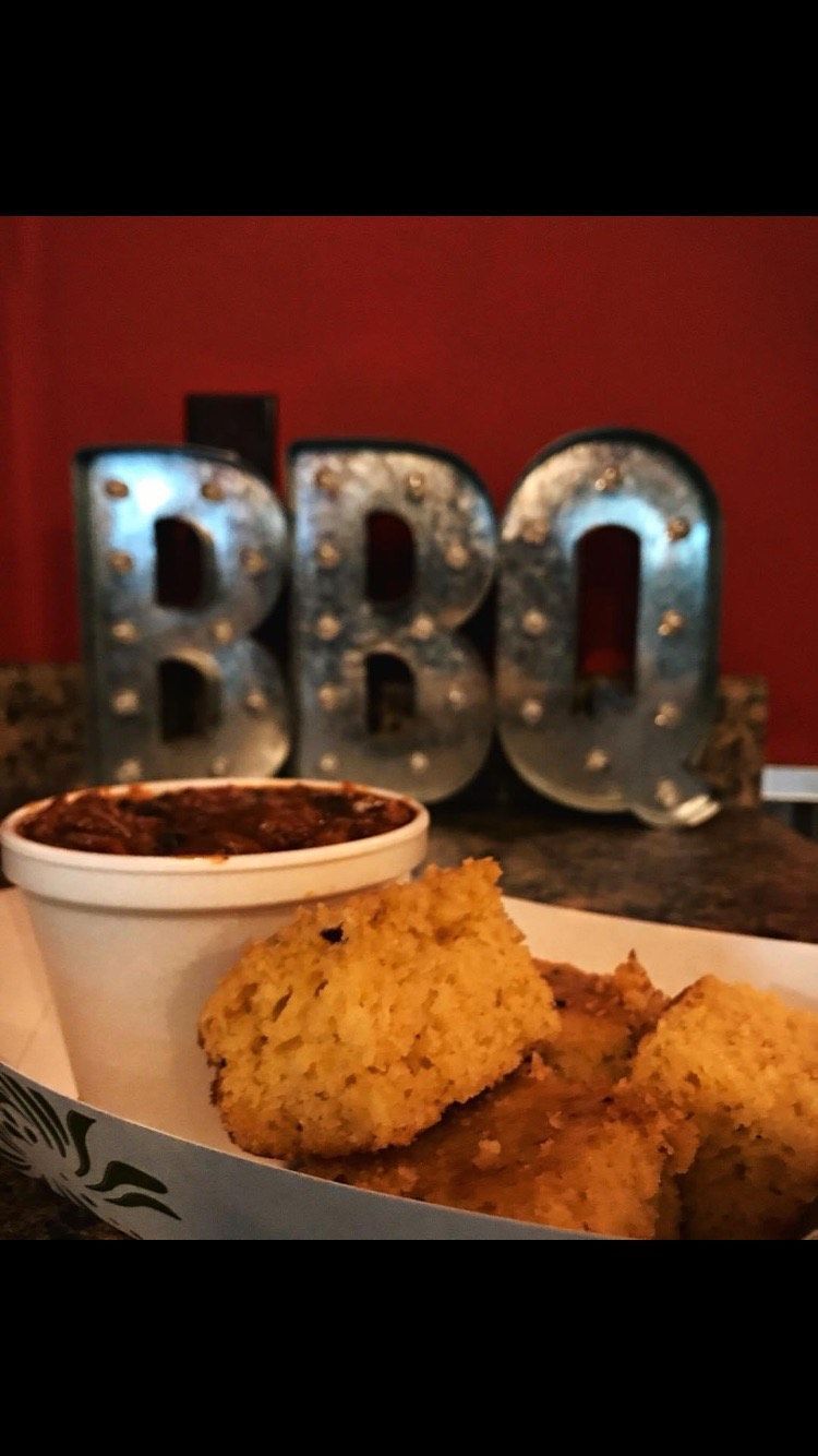A paper tray with a cup of pulled meat and pieces of cornbread, set against a red wall with metal 