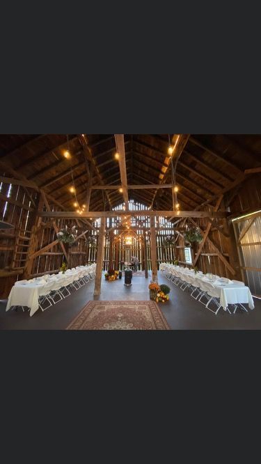 An indoor rustic barn wedding venue featuring two long tables set with white tablecloths and warm string lighting overhead.