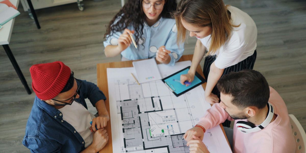 Four people collaborate around a wooden table, reviewing a large floor plan blueprint and a tablet.