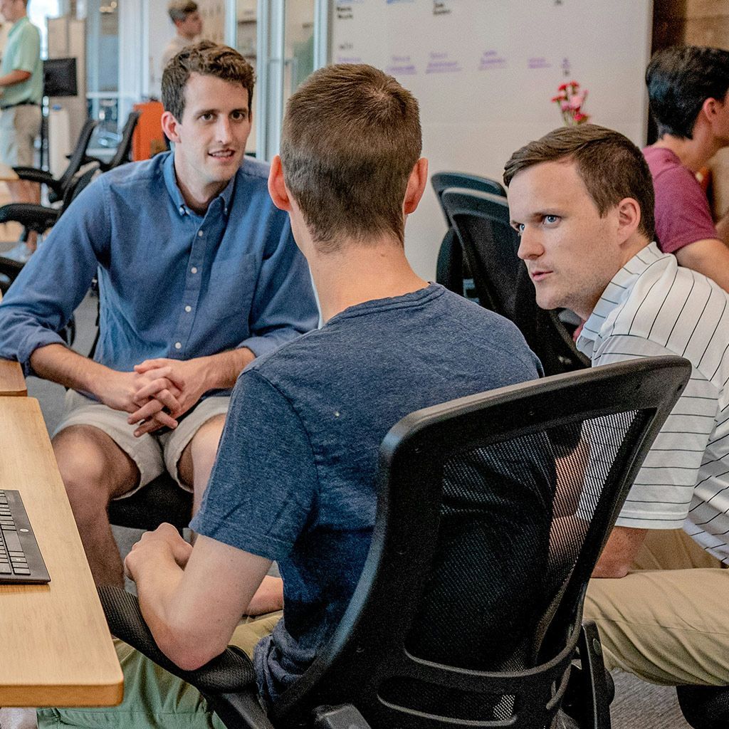 Three people sit in an office, engaged in a discussion while facing one another across a desk.