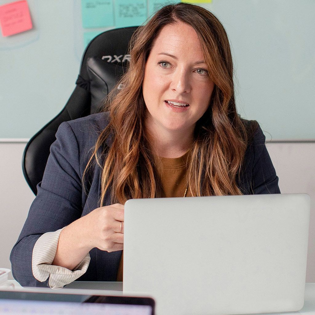 A professional sitting at a desk with a laptop, speaking with an engaged expression in an office with whiteboards.