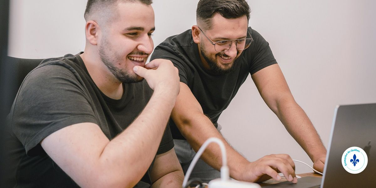 Two people in dark t-shirts look at a laptop screen together while smiling in an office setting.