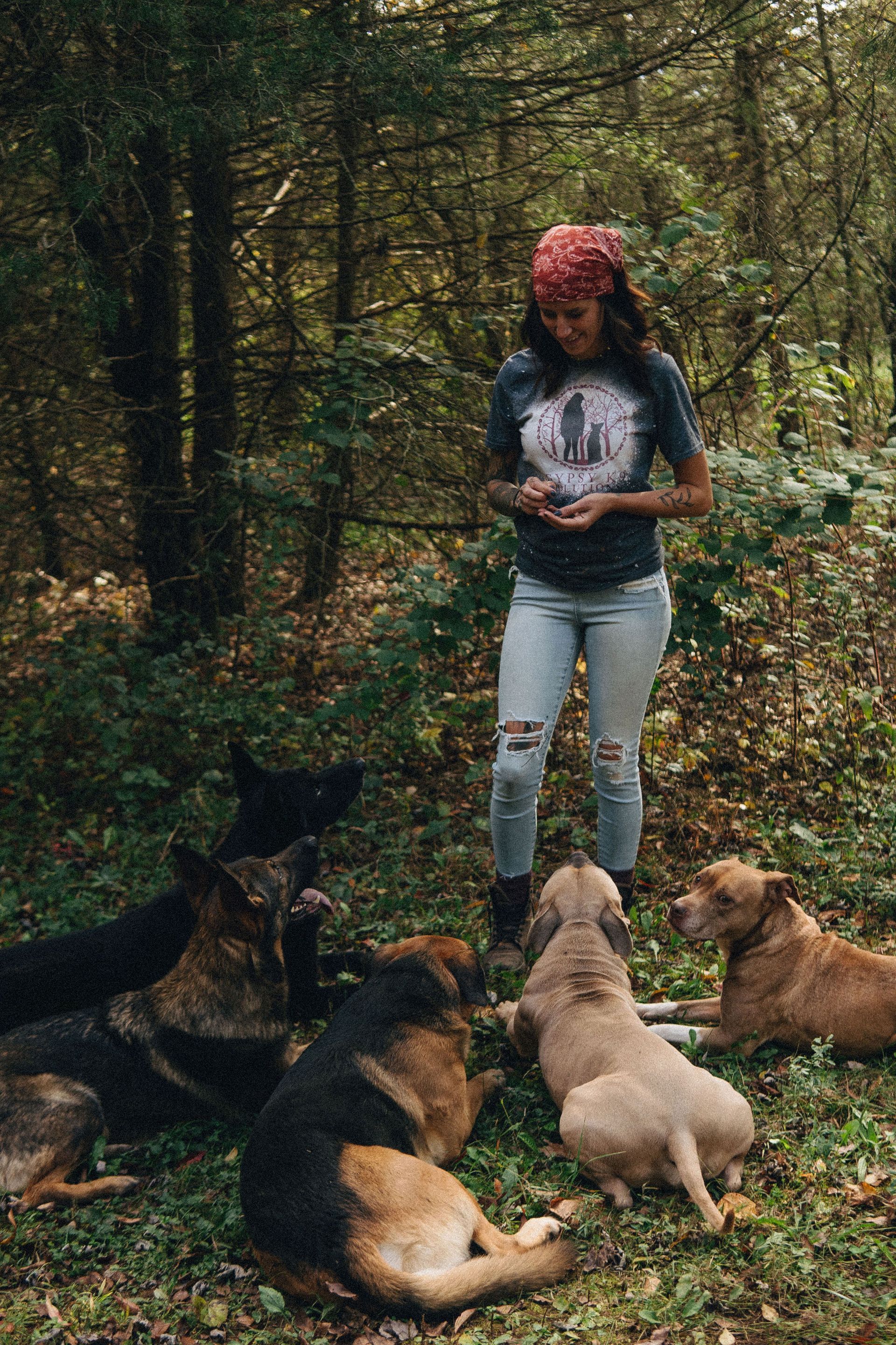 Woman in jeans and bandana stands with several dogs in a forest.