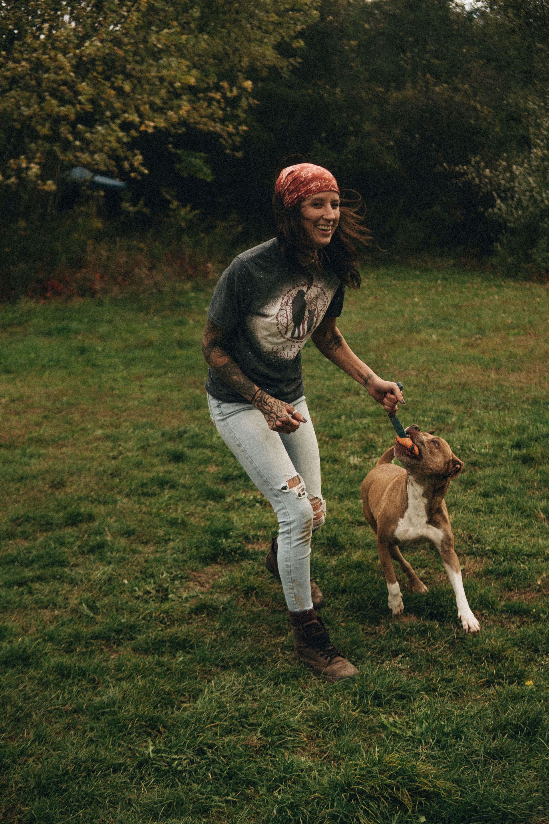 Woman in jeans and bandana playing with a dog on grass, pulling a leash.