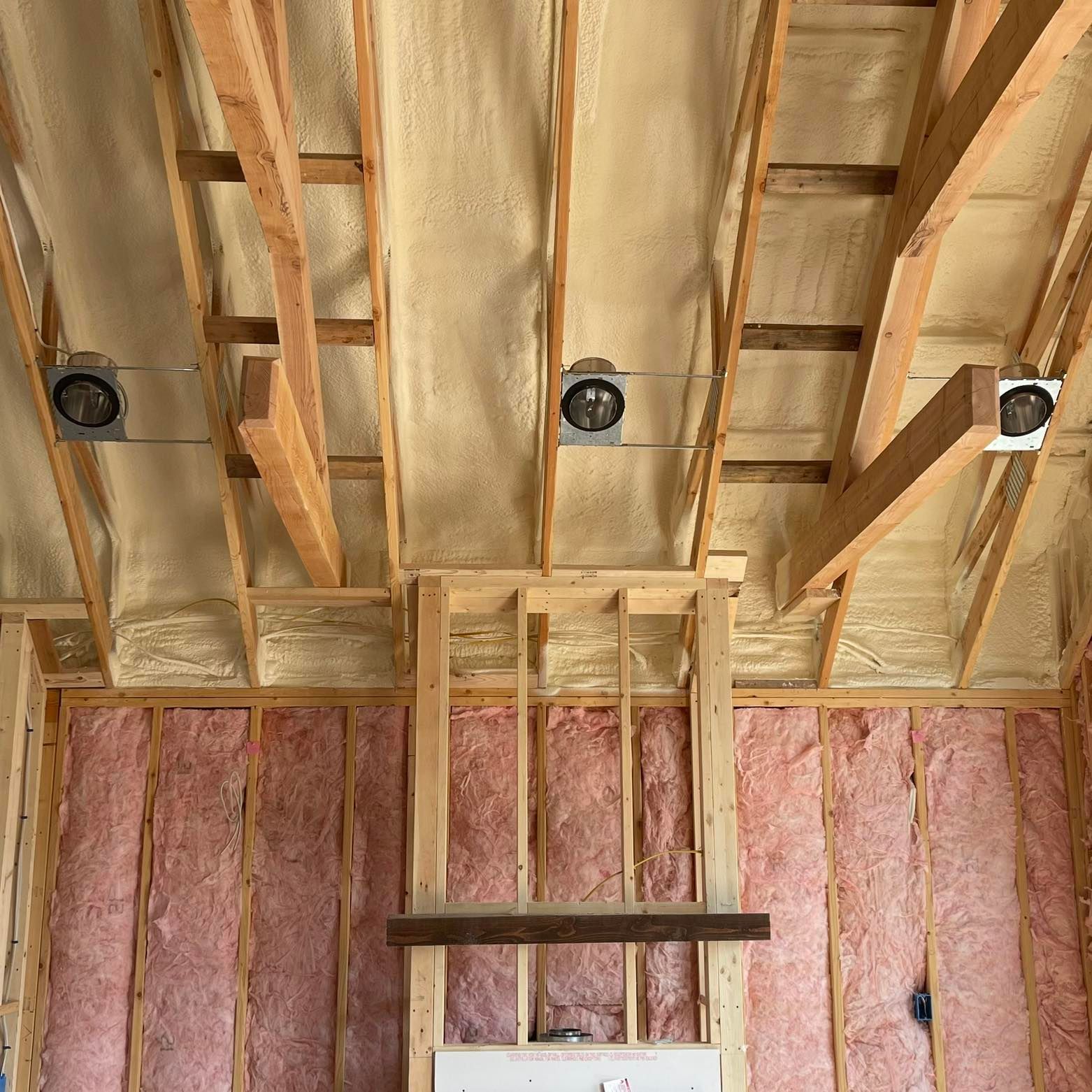 Unfinished ceiling and wall framing with yellow spray foam insulation and pink fiberglass batts, including light housings.