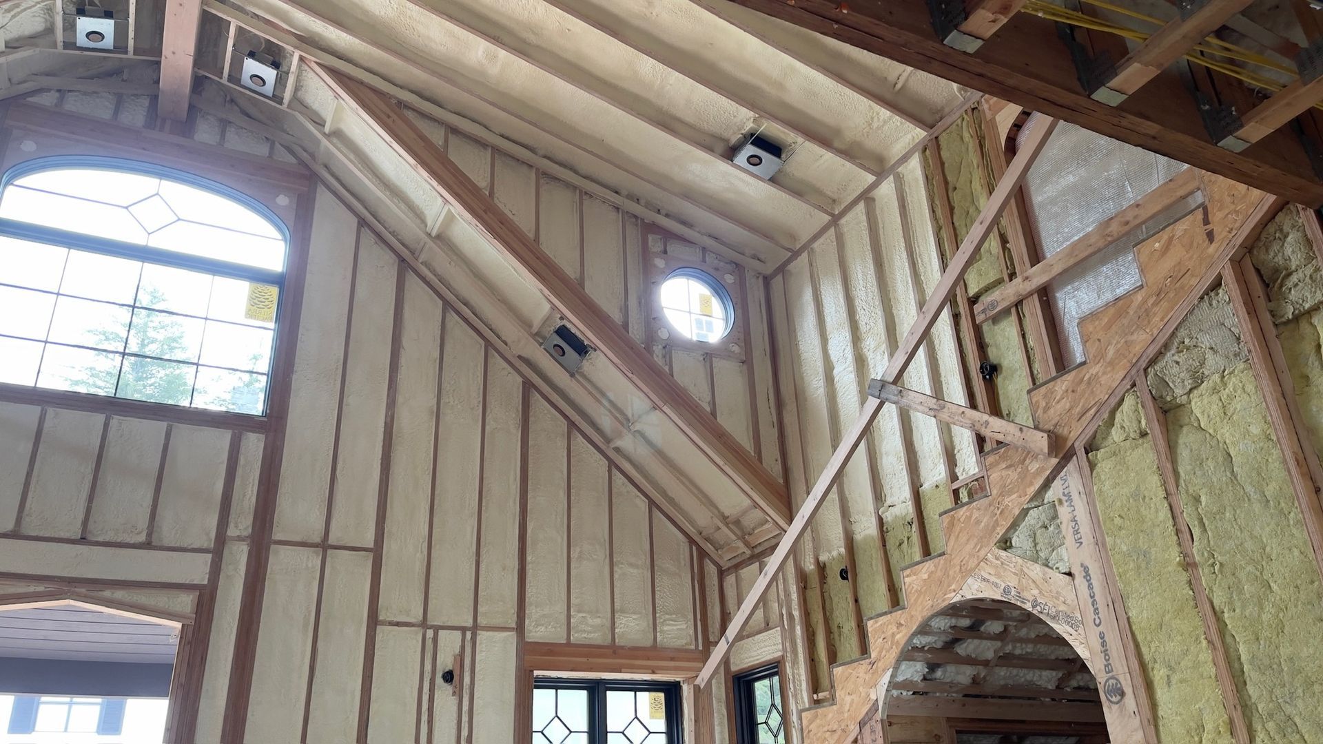 Interior view of a room under construction with exposed wooden rafters, wall studs, spray foam insulation, and windows.