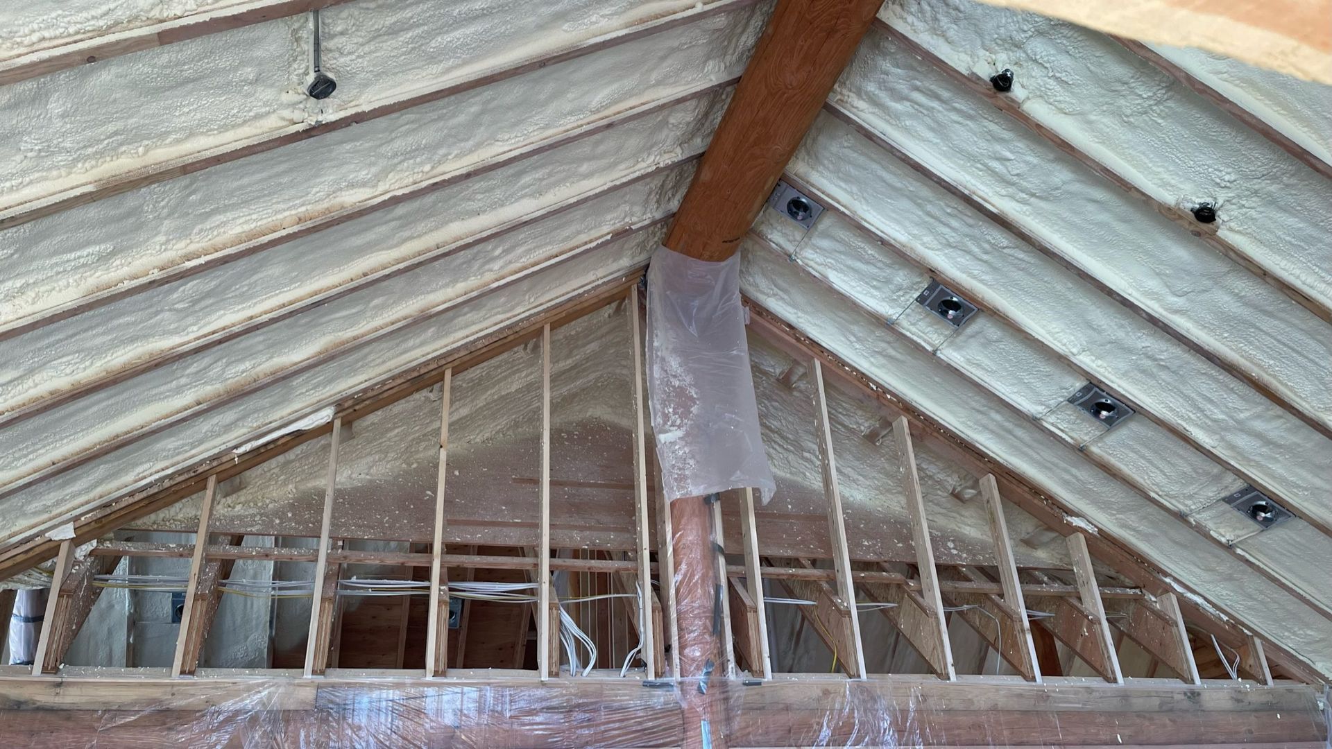 Attic rafters and roof joists insulated with spray foam, featuring a wooden ridge beam and a plastic-covered center post.