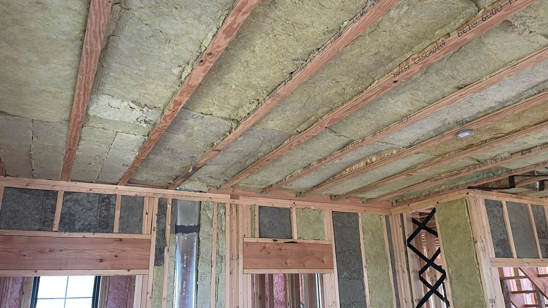 Interior view of a residential construction site featuring wood framing and green insulation batts in the ceiling and walls.