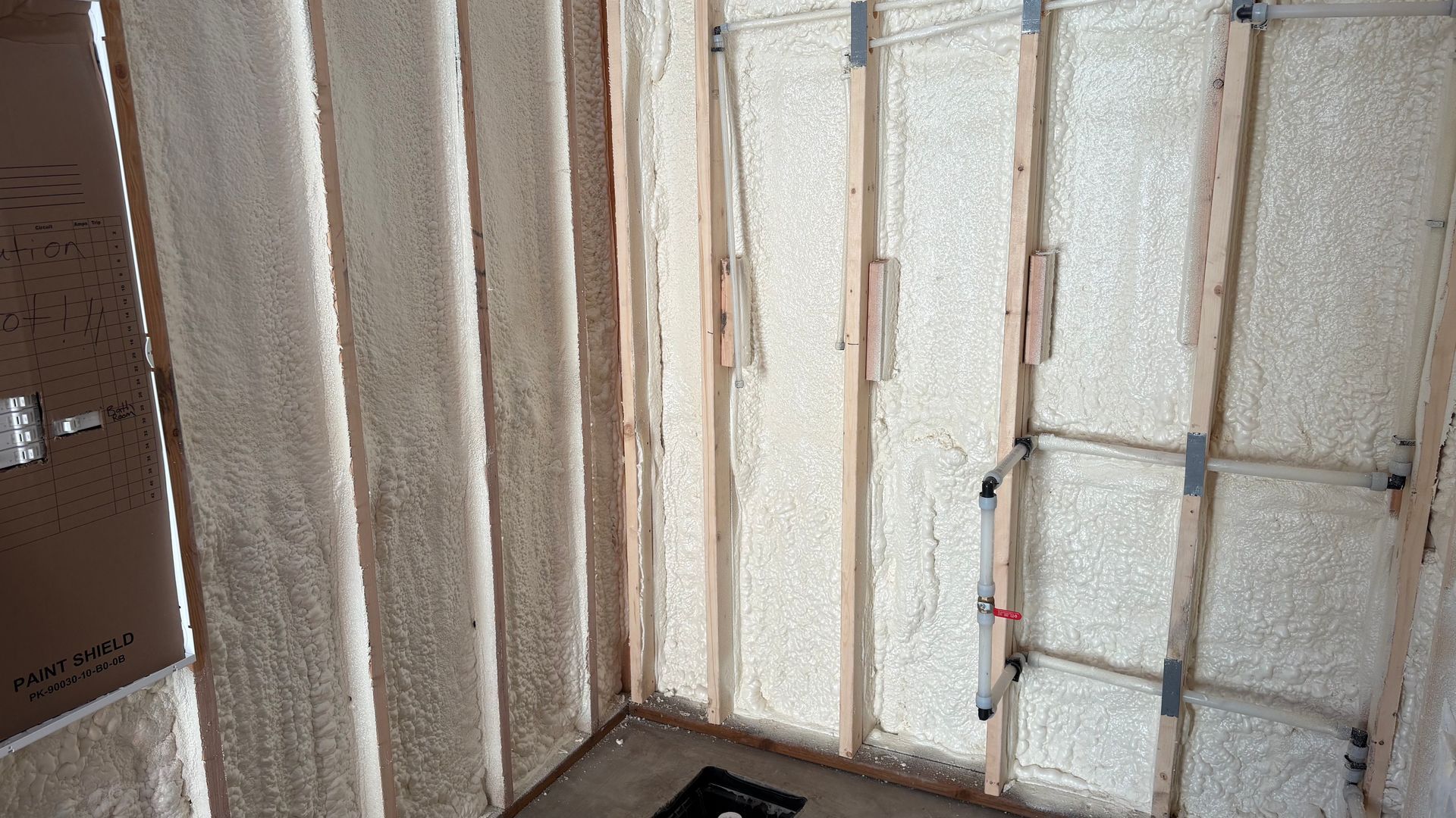 A wall with exposed wooden studs insulated with cream-colored spray foam, showing some plumbing pipes and electrical wire.