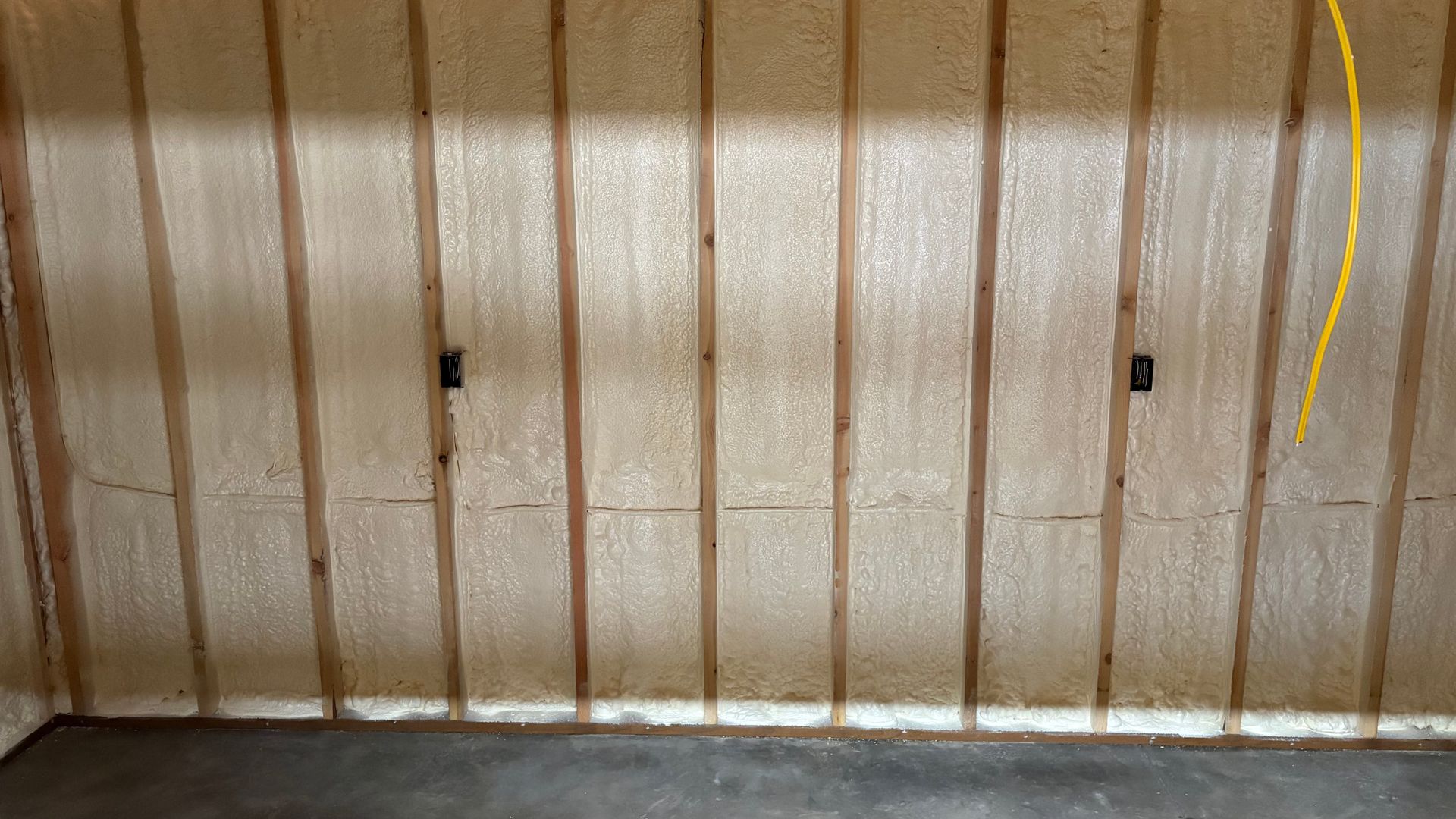 Spray foam insulation installed between wooden wall studs in a garage with two electrical outlets and a hanging cable.