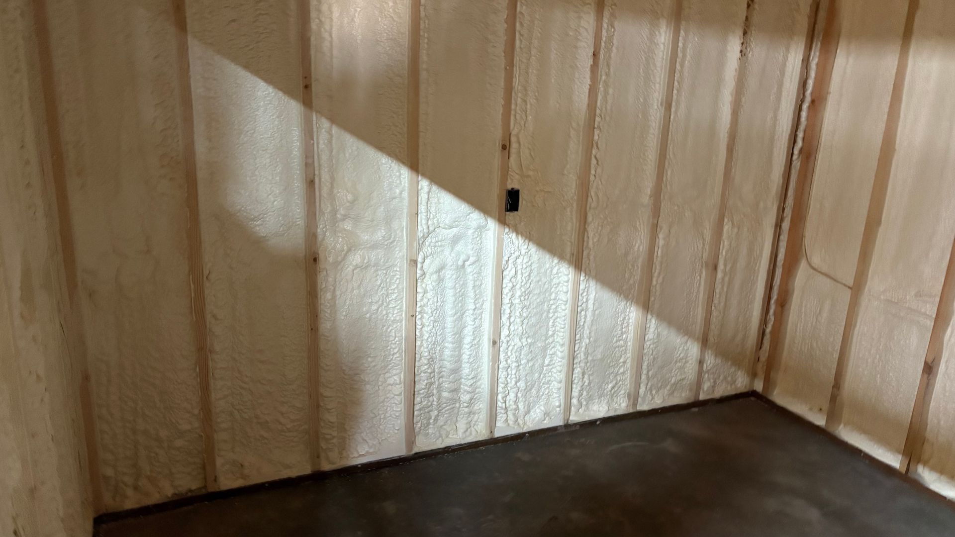 Interior wall framing and concrete floor, with white spray foam insulation filling the stud cavities.