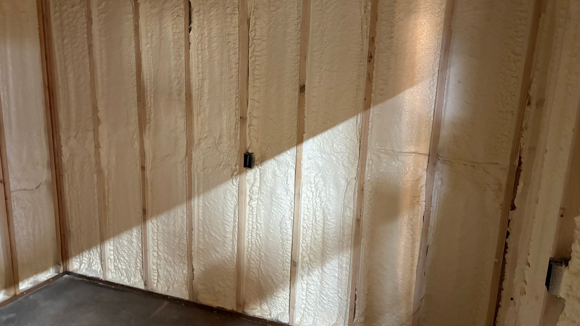 An unfinished room with wooden wall studs filled with light-colored spray foam insulation and a single electrical box.