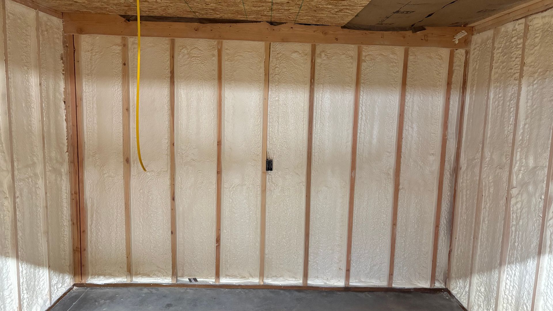 Interior wall frame with spray foam insulation between wooden studs, featuring a yellow wire and electrical outlet.