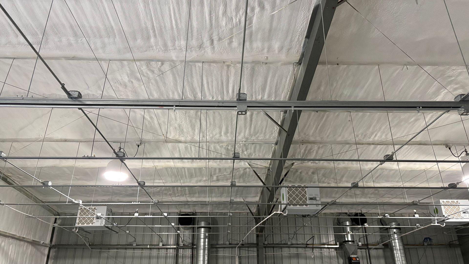 An interior view of a commercial ceiling with white spray foam insulation, metal framing, electrical conduits, and HVAC.