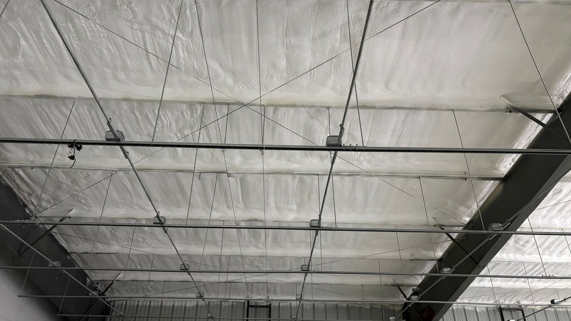 Underside of a metal industrial roof covered in thick white spray foam insulation, with support beams and wiring visible.