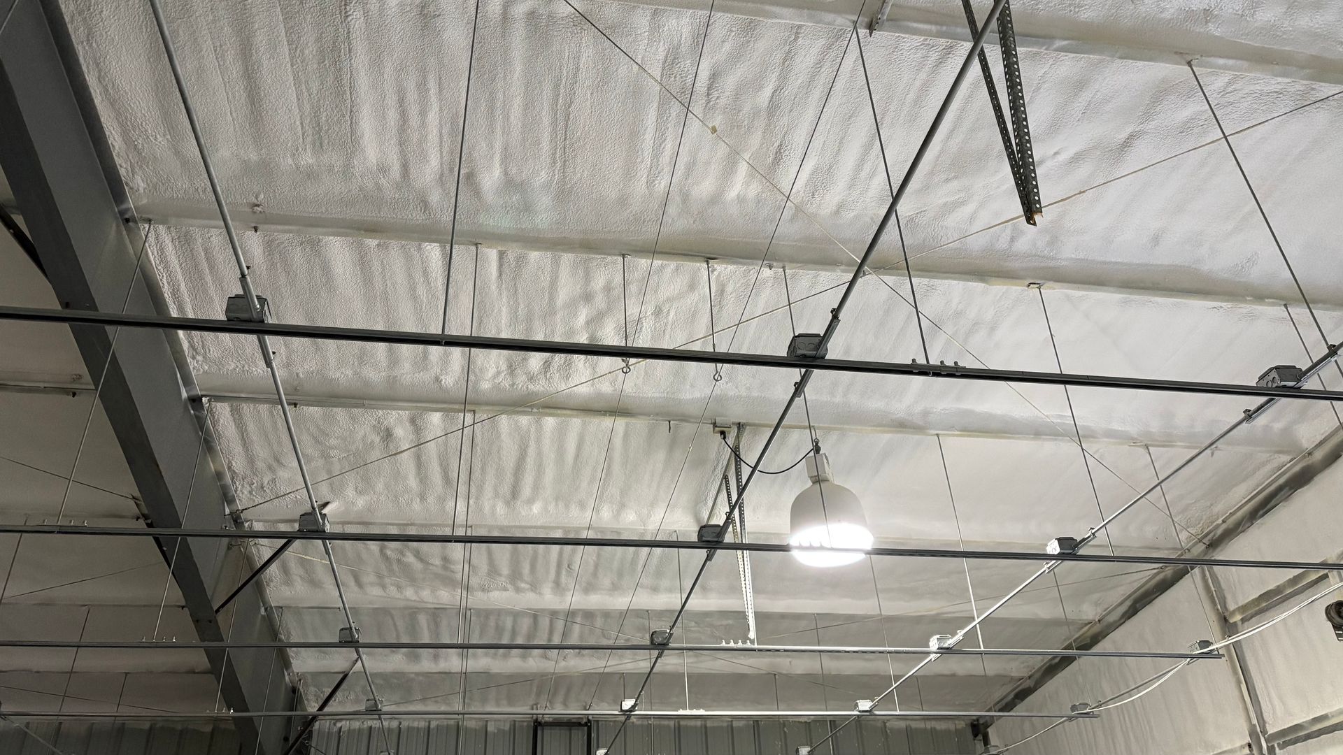 An industrial ceiling with white spray foam insulation, steel beams, and hanging electrical lighting fixtures.