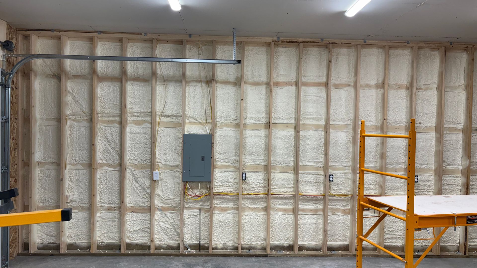 An interior wall with wooden framing insulated with white spray foam, featuring an electrical panel and scaffolding.