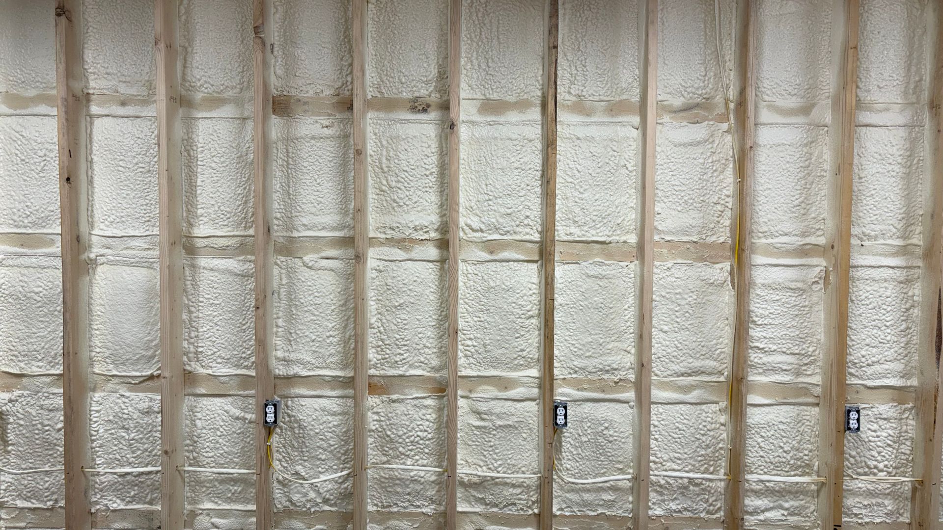 Spray foam insulation installed between wooden wall studs with three electrical boxes visible.