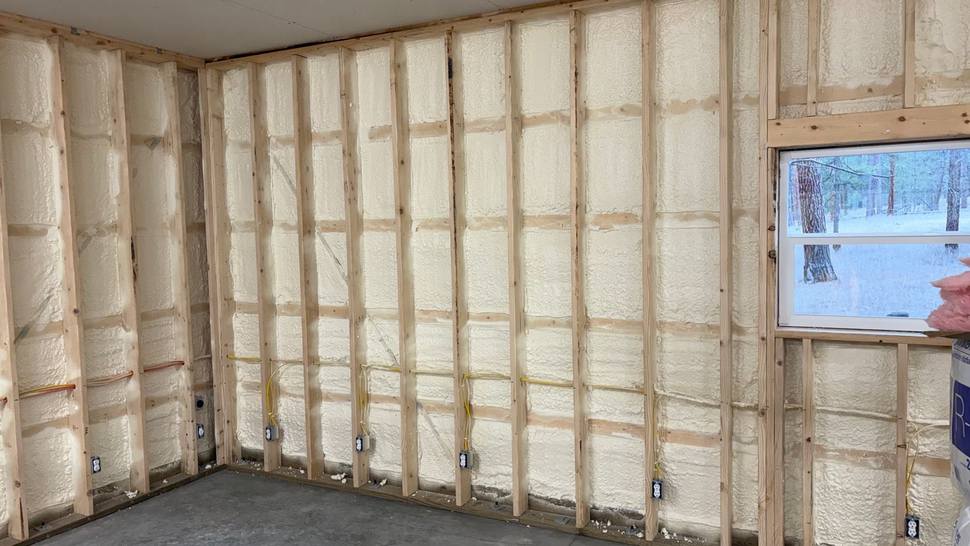 An interior view of a room under construction with wood wall framing filled with white spray foam insulation.