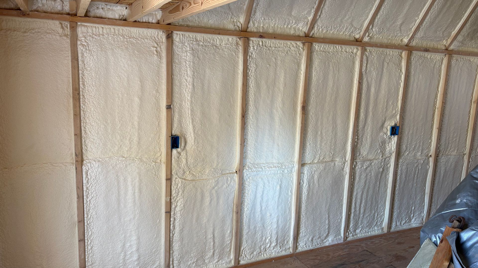 Spray foam insulation installed between the wooden wall studs and rafters of an unfinished interior space.