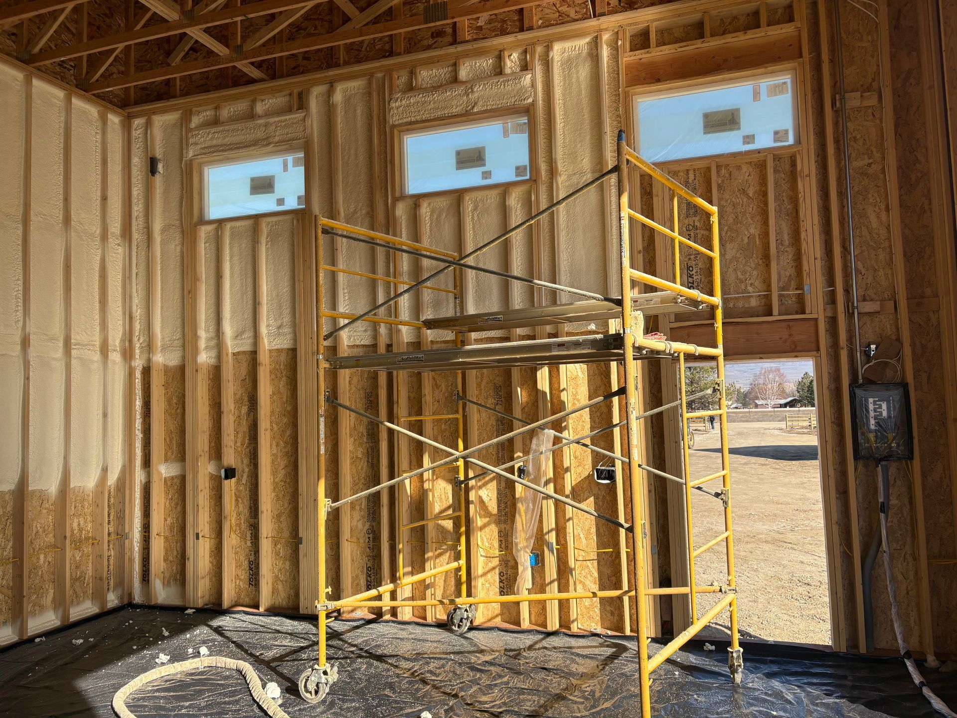 Yellow metal scaffolding stands inside a partially built room with wood-framed walls and three rectangular windows.