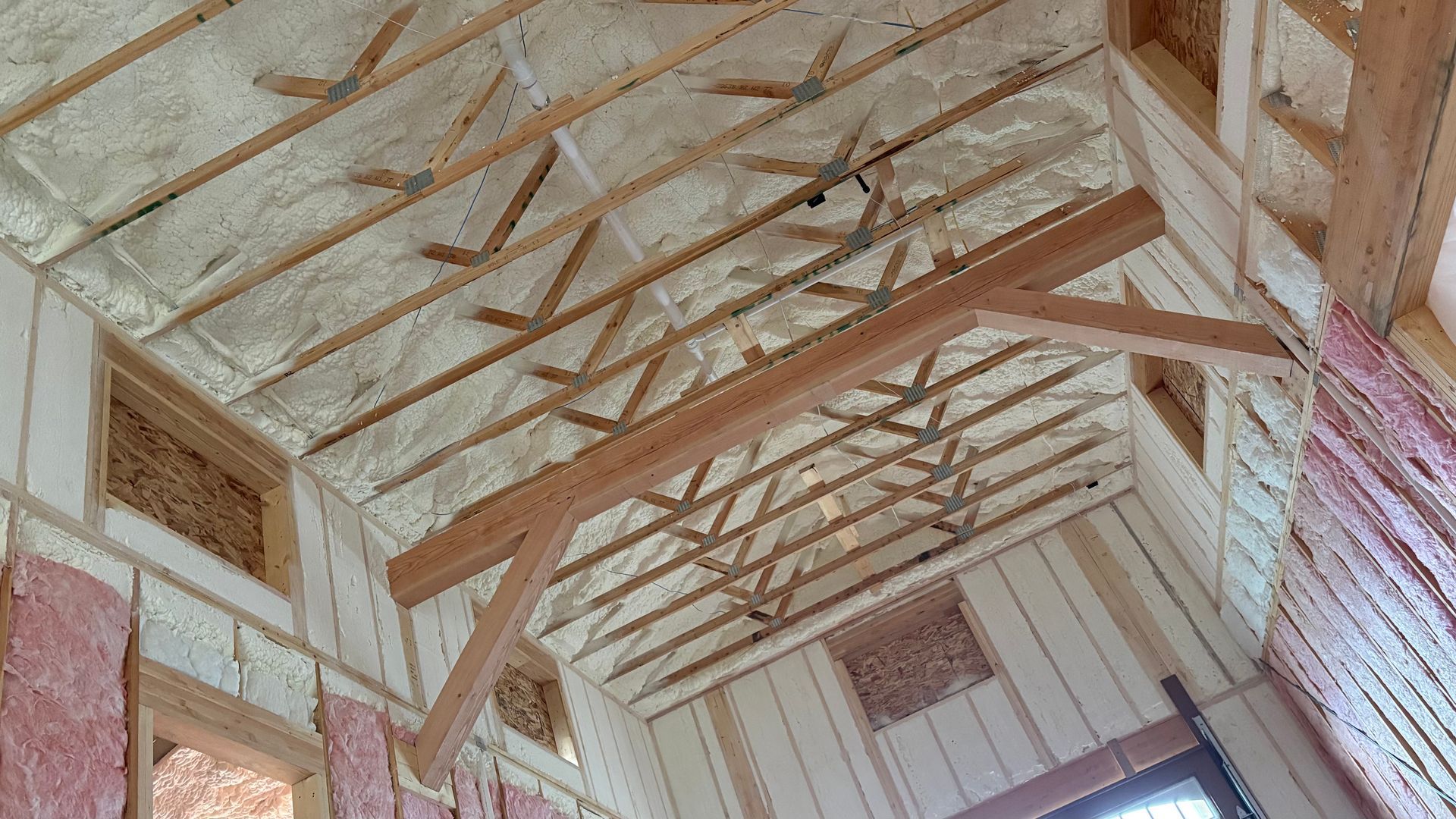 An interior view of a room under construction, showing wooden ceiling trusses, exposed insulation, and a timber beam.