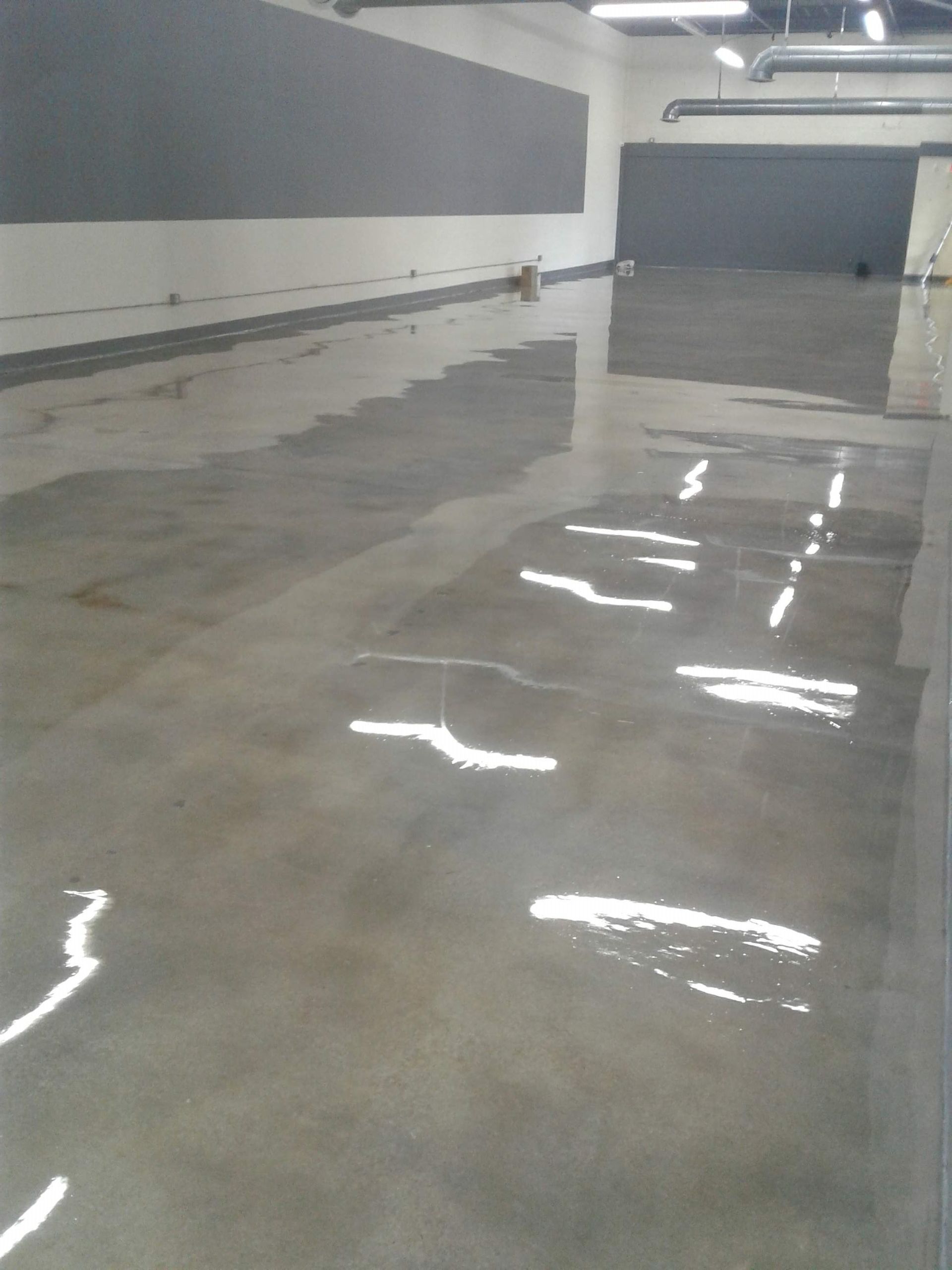 Empty indoor garage with glossy wet concrete floor and gray walls, reflecting overhead lights.