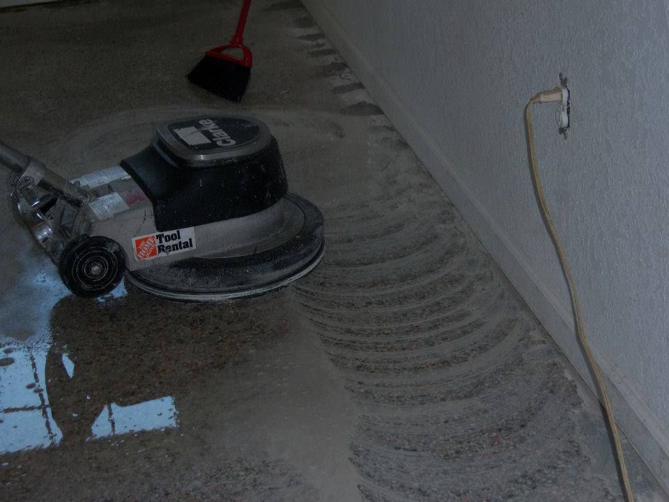 Floor buffer cleaning a wet concrete floor beside a wall and power cord.