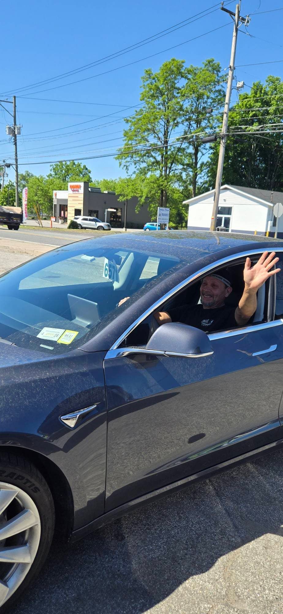 Person waving from the driver’s seat of a dark car on a sunny street