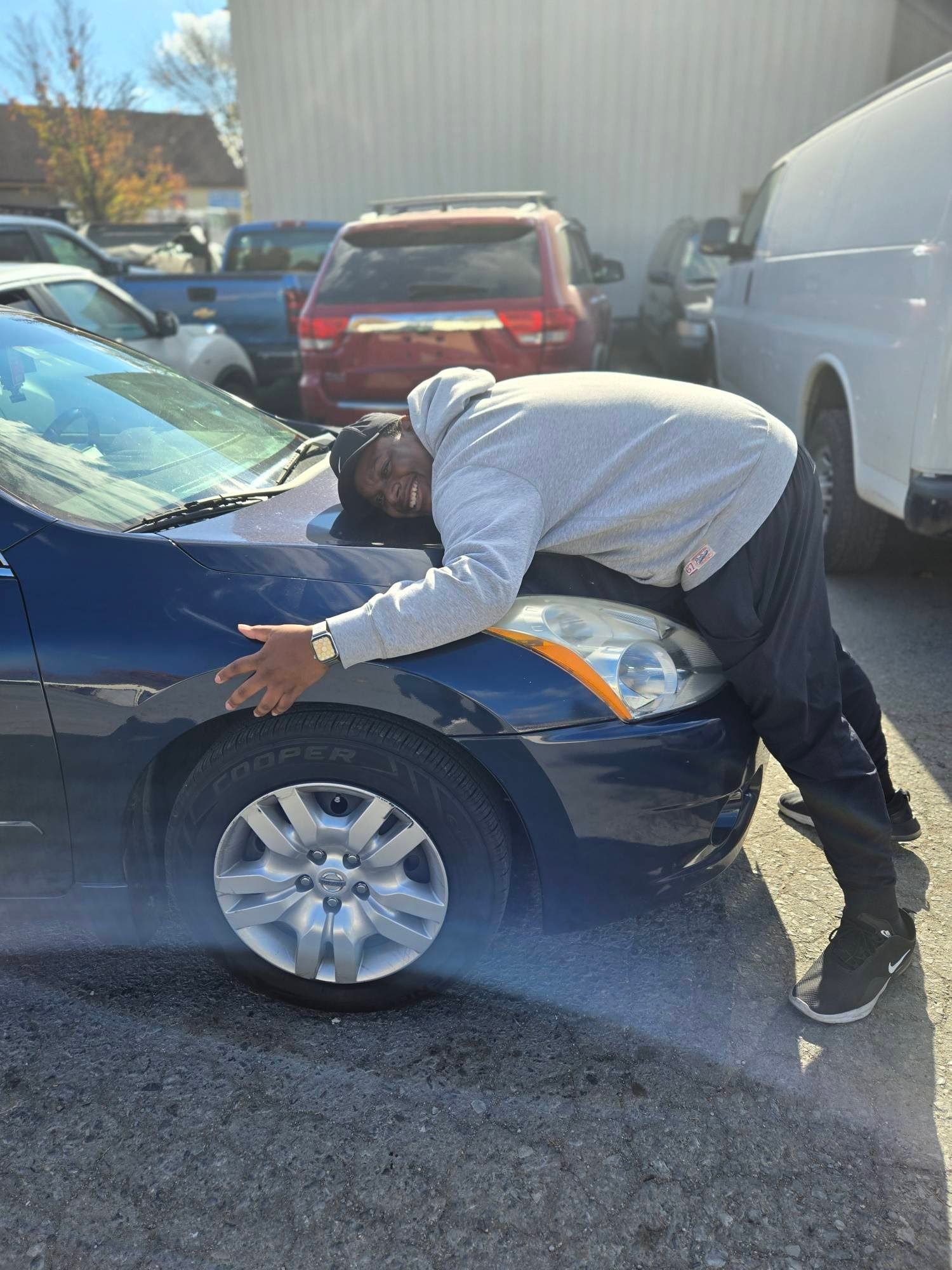 Person leaning into the hood of a dark blue car in a parking lot, with a red SUV and white van nearby.