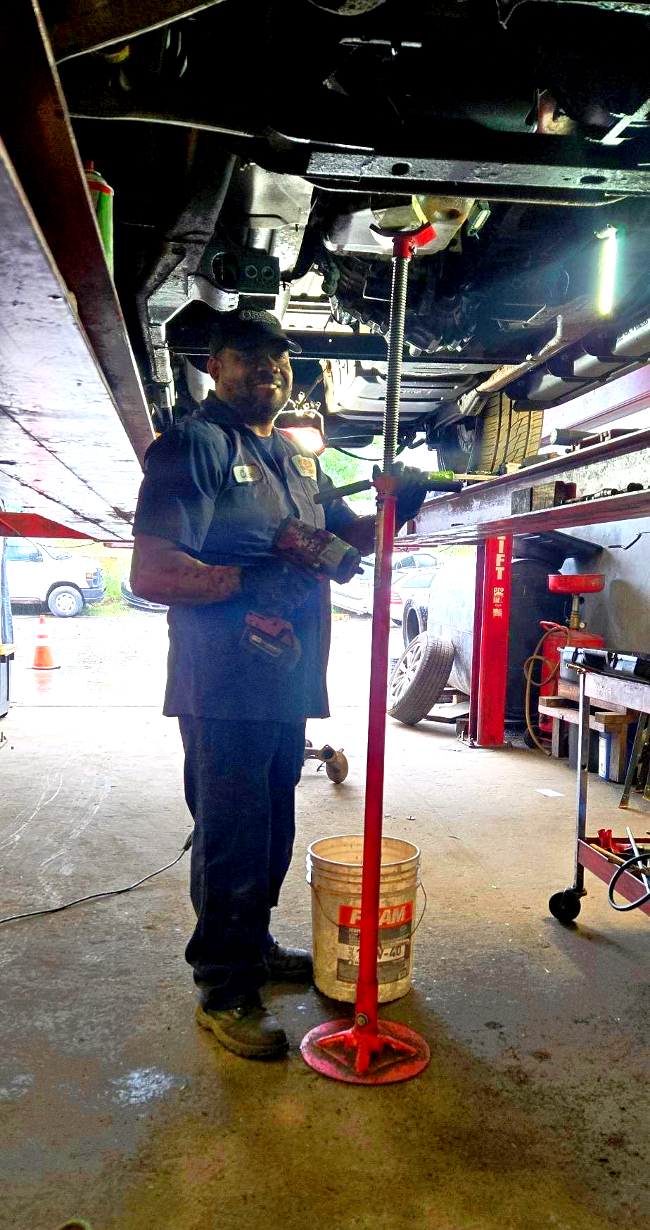 Mechanic standing under a car on a hydraulic lift in a garage, working near a red support stand.