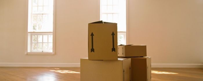 Stacked cardboard moving boxes in a sunlit empty room with windows.