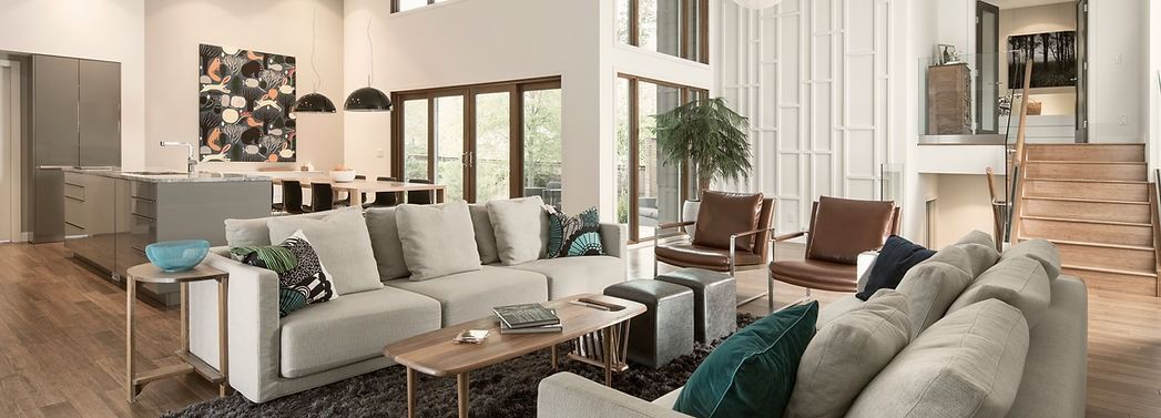 Modern open-plan living room with beige sofas, green cushions, armchairs, staircase, and dining area
