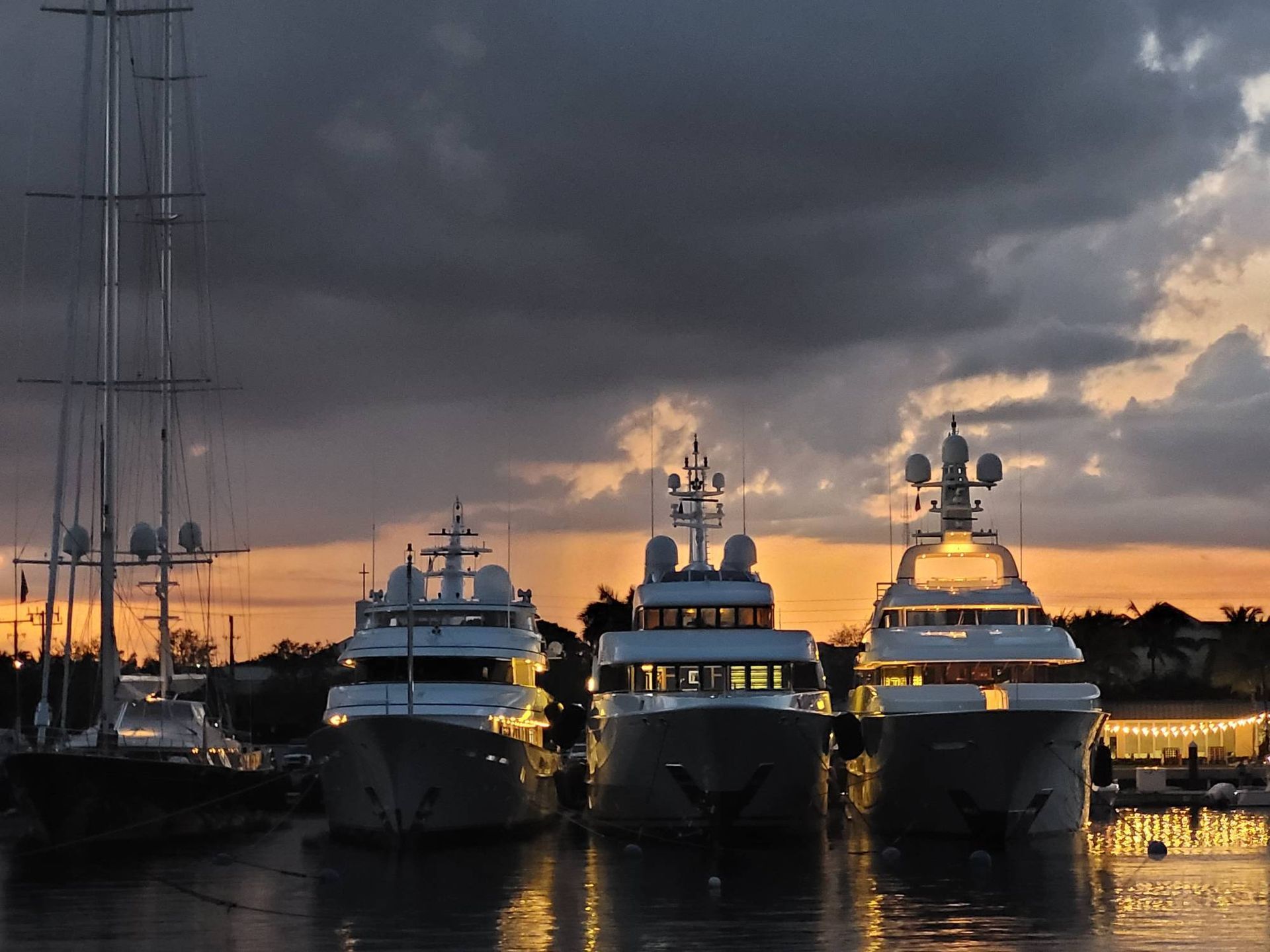 Three large luxury yachts docked at a marina during a cloudy sunset, their lights reflecting on the water.
