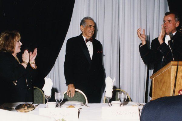 A man in a tuxedo stands at a podium with two other men applauding