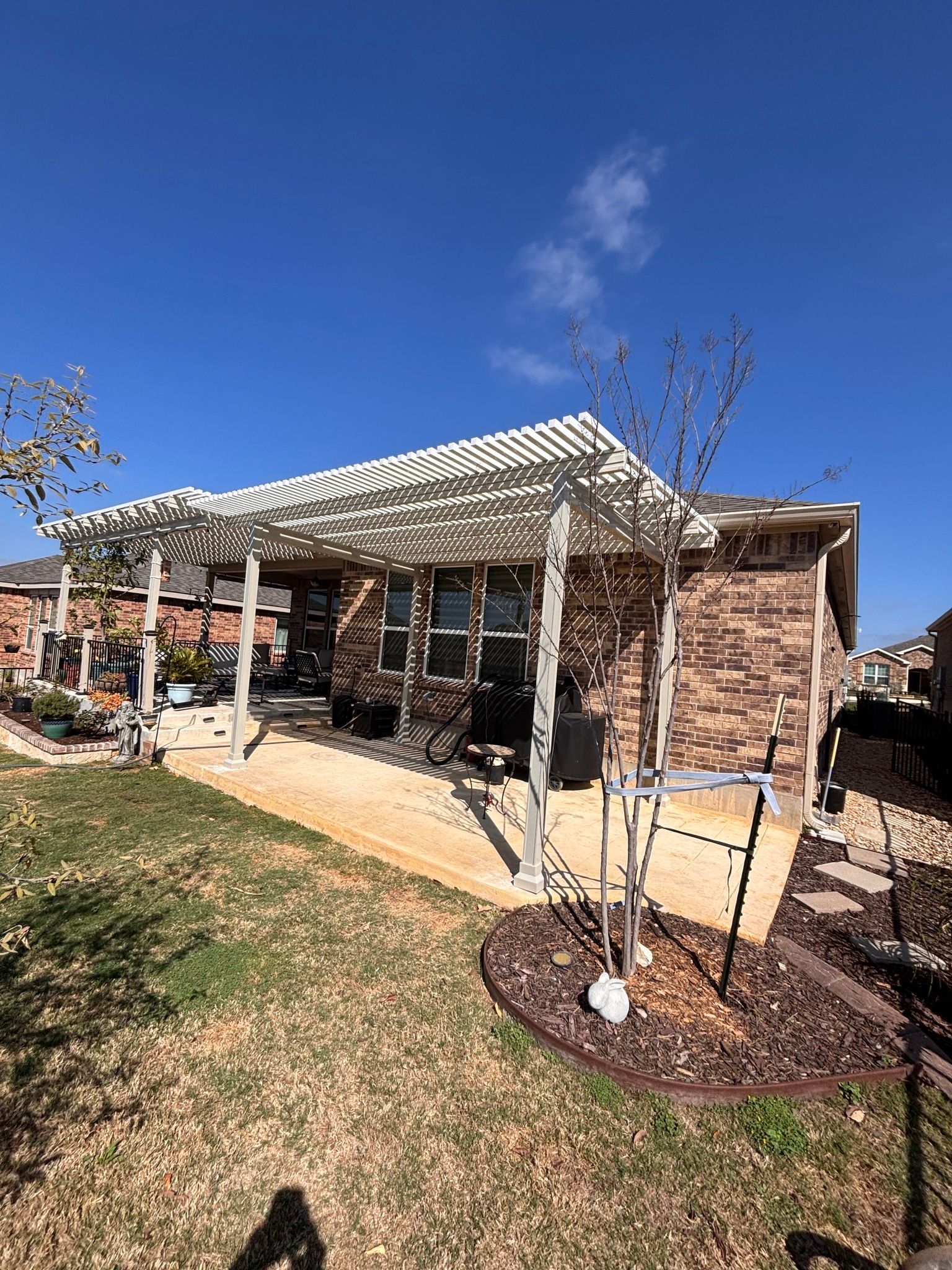 A brick house patio features a white wooden pergola, a stone-colored concrete floor, and a small yard under a blue sky.