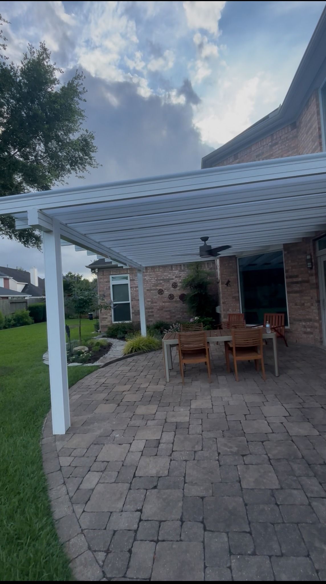 A patio with a white pergola, table, and chairs sits on a paved stone surface attached to the back of a brick house.