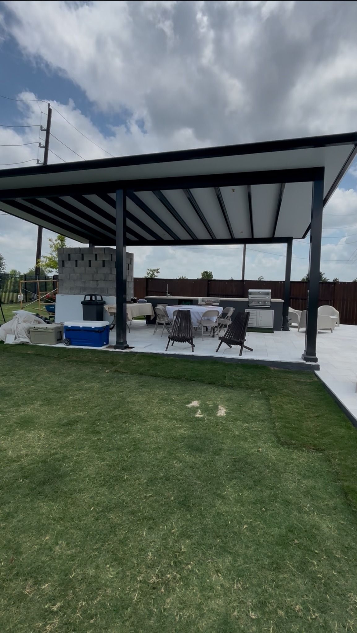 An outdoor patio area with a black-framed roof covering a concrete space with a grill, outdoor furniture, and a cooler.