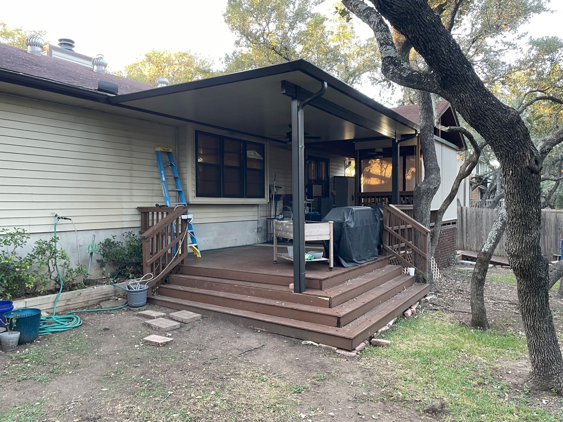 A wooden deck with a covered roof, stairs, and railings attached to the side of a light-colored house near large trees.