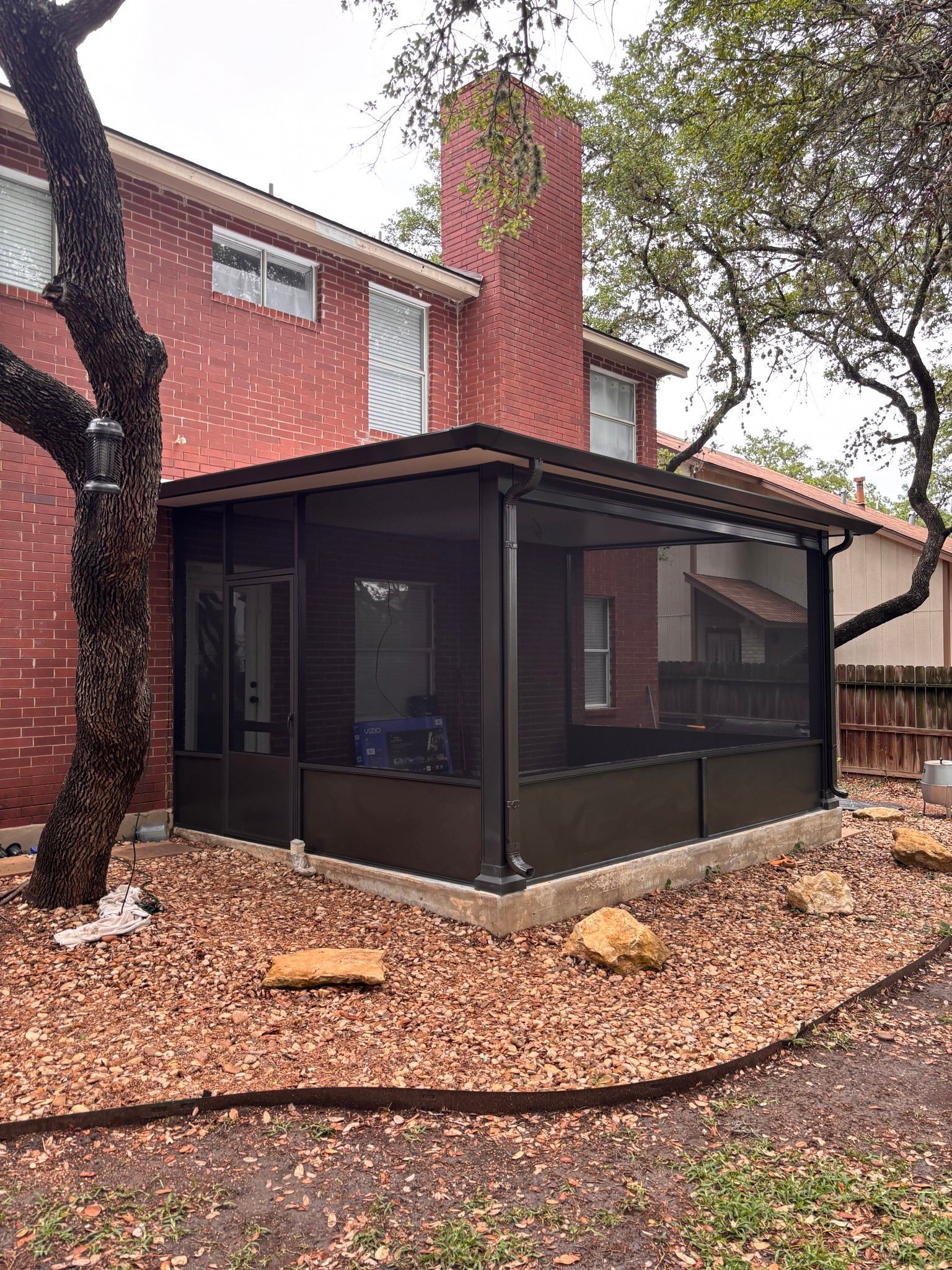 A screened-in porch with a dark frame attached to the side of a red brick house with a brick chimney and a gravel yard.